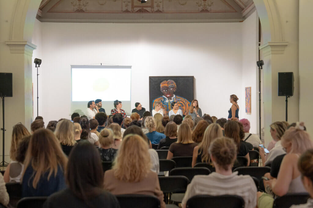 Horizontal photograph. Audience seated in a bright exhibition space before a panel discussion. Several people are discussing on a stage, behind them are a work of art and a projection screen.